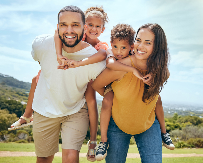 Smiling parents giving their two children piggyback rides outdoors on a sunny day