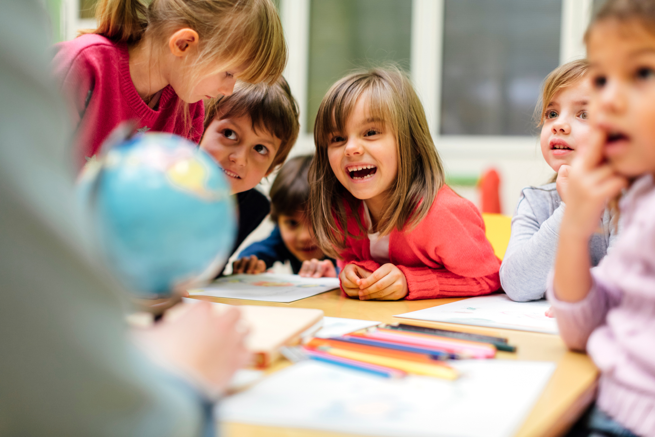 Preschool children gathered around a table, laughing as they look at a globe with their educator